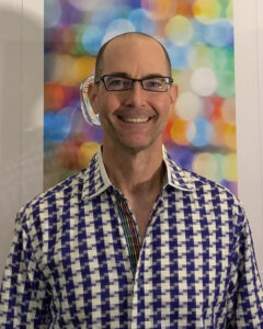 A man wearing glasses and a patterned shirt stands in front of a colorful, abstract background with circular light shapes, reflecting the creative spirit of Ars Nova Singers’ Community-Driven approach.