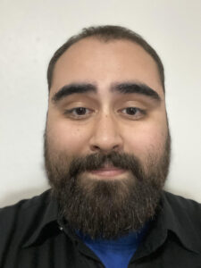 A bearded man with dark hair and eyebrows, reminiscent of Joseph Berrios, is wearing a black shirt and looking directly at the camera against a plain light background.