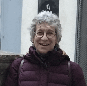 Elissa Guralnick, an older woman with short gray hair and glasses, smiles at the camera in a maroon jacket. The background features a light wall and part of a blue door.