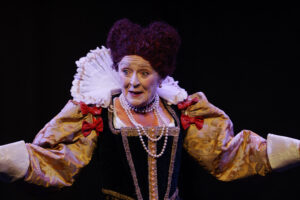 Person dressed in an elaborate historical costume with puffed sleeves, lace ruff, pearls, and red bows—evoking the grandeur of Queen Elizabeth I—performs on stage against a dark background.
