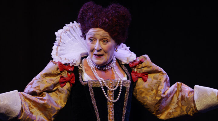 Person dressed in an elaborate historical costume with puffed sleeves, lace ruff, pearls, and red bows—evoking the grandeur of Queen Elizabeth I—performs on stage against a dark background.