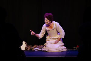 A person in historical costume kneels on stage, pointing forward with an expressive face against a dark background, evoking the grandeur of Queen Elizabeth I's era.