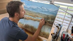 A person is painting a landscape scene on a canvas—mountains, trees, and water—at a desk with art supplies by a window, capturing the spirit of Shared Visions 2026.