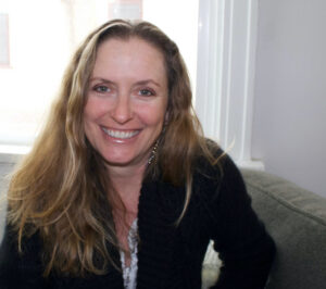 A woman with long, light brown hair and a black sweater smiles while sitting on a gray couch near a window, embodying the spirit of Shared Visions 2026.