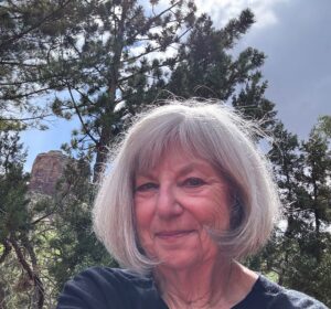 An older woman with gray hair smiles outdoors, enjoying the beauty of pine trees and a rock formation beneath a partly cloudy sky—capturing the spirit of Shared Visions 2026.