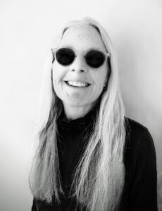 A woman with long gray hair wears round sunglasses and a dark top, smiling in front of a plain light background, embodying the spirit of Shared Visions 2026.
