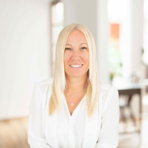 A woman with long blonde hair wearing a white blouse smiles at the camera in a bright, modern indoor setting, capturing the spirit of Shared Visions 2026.