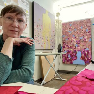 A woman with short hair and glasses sits at a table with pink fabric in front of her; abstract artwork and a skeleton model in the background hint at the creative spirit of Shared Visions 2026.
