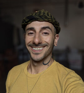 A person wearing a yellow shirt and patterned headwrap smiles at the camera in an indoor setting with a blurred background.
