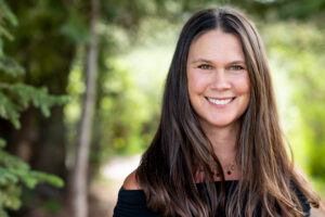 Woman with long brown hair smiling outdoors, wearing a black off-the-shoulder top and a necklace, with greenery in the blurred background.
