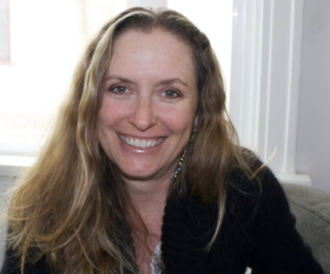 A woman with long, light brown hair and a black sweater is smiling while sitting indoors near a window, her expression thoughtful as if lost in reflections in water.