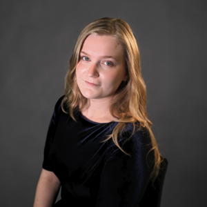 A woman with long blonde hair and a neutral expression sits against a plain dark background, wearing a black top—capturing the understated elegance of Shared Visions 2026.