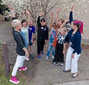 A group of children and two adults stand outdoors near a blooming tree in EDEN Colorado, all raising one arm and smiling during an activity.
