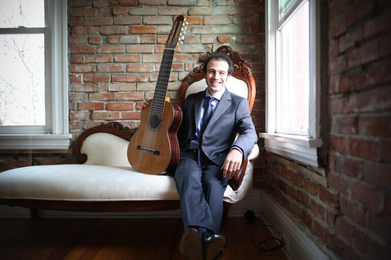 Guitarist Nicolò Spera seated on a settee beside his ten-string classical guitar in a brick-walled room