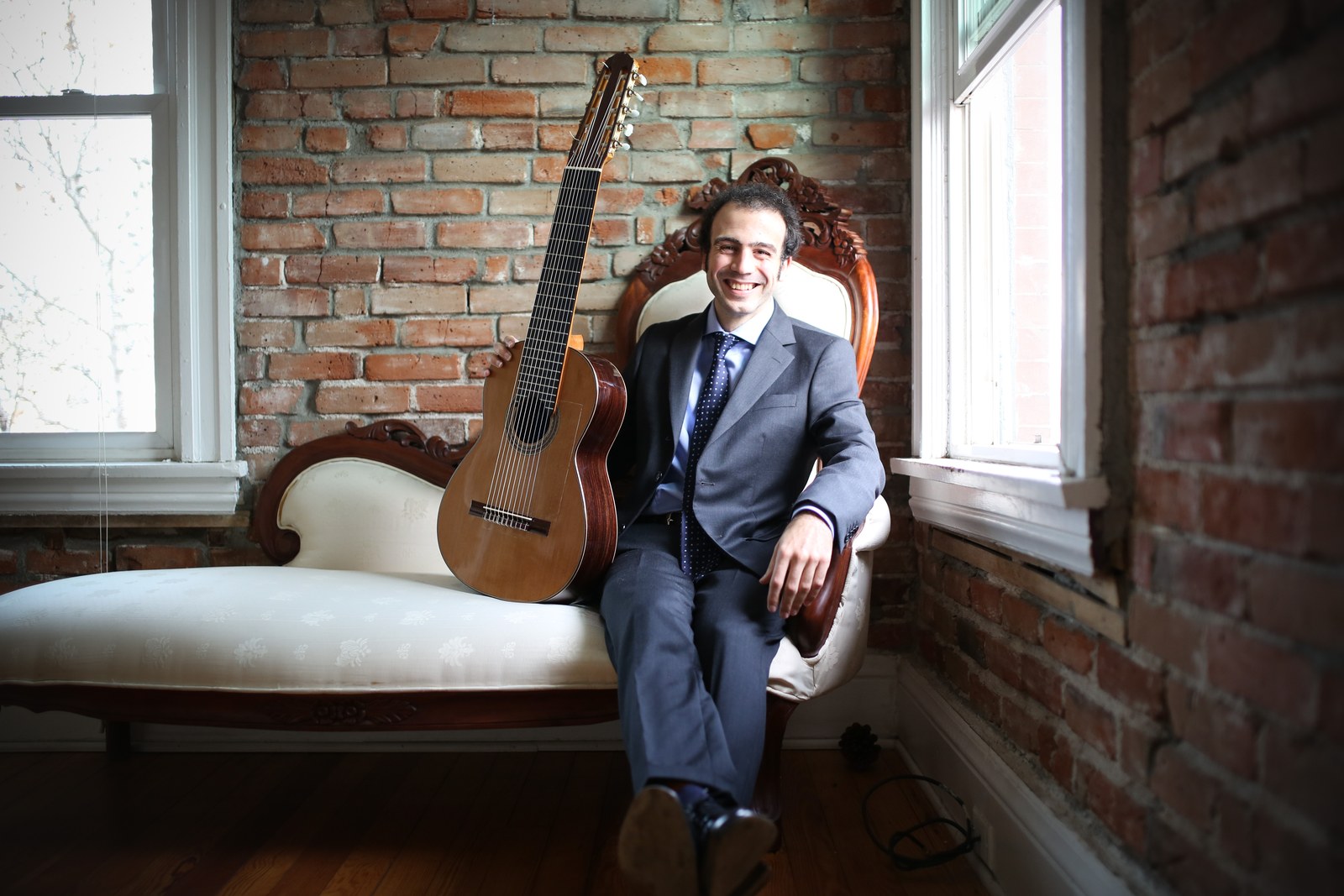 Guitarist Nicolò Spera seated on a settee beside his ten-string classical guitar in a brick-walled room