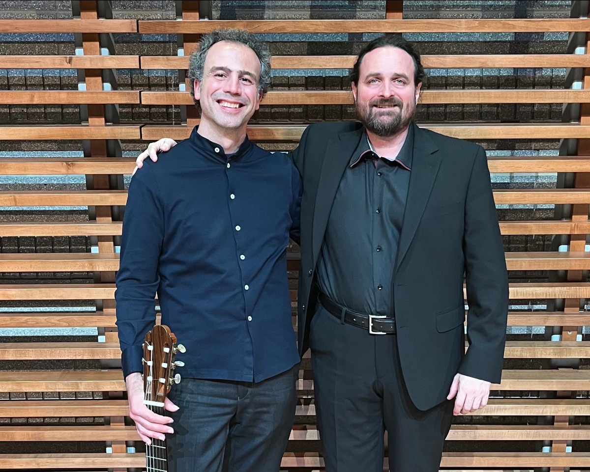 Guitarist Nicolò Spera holding his classical guitar, standing beside composer Corrado Margutti in a concert hall