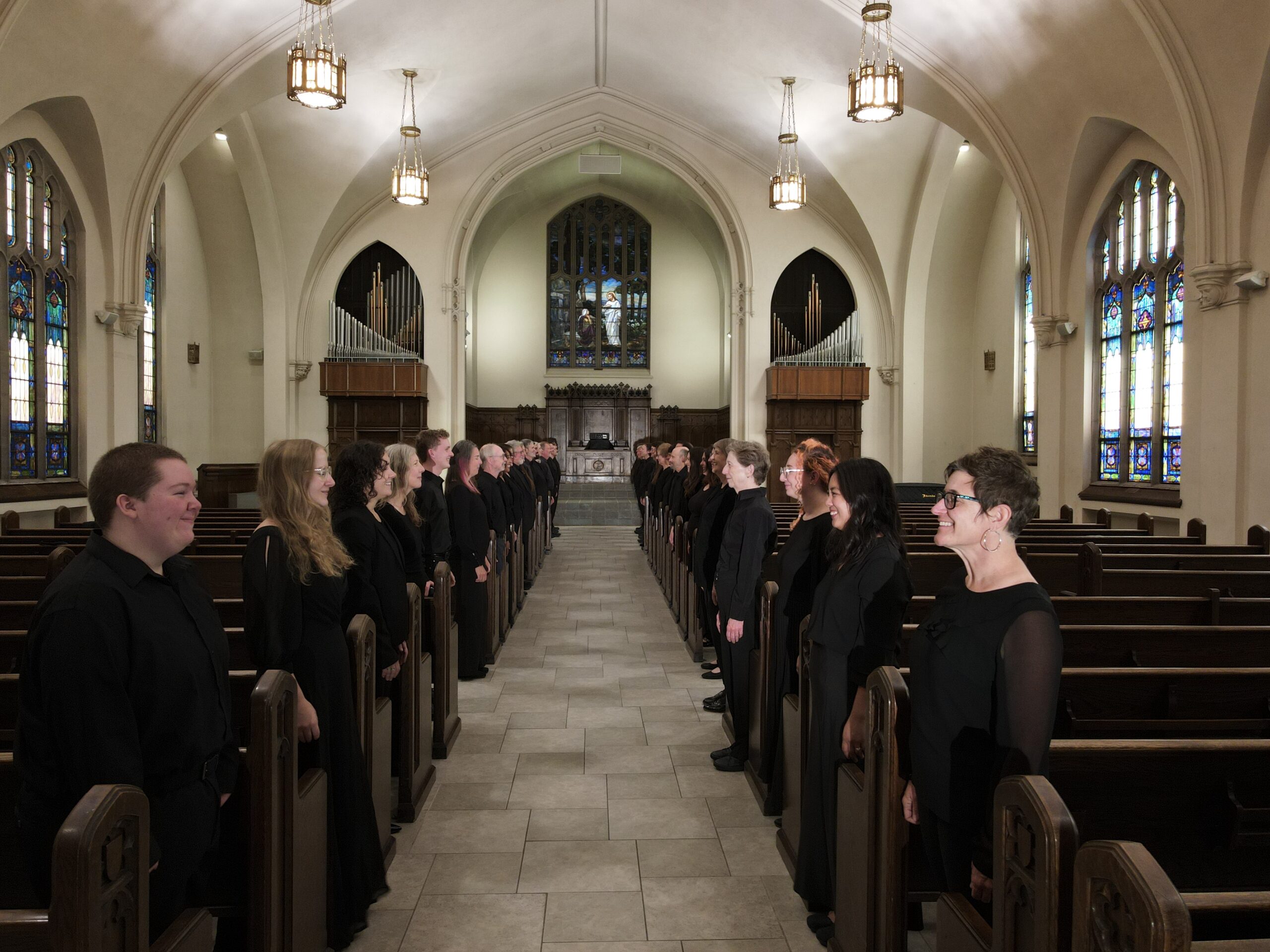 Ars Nova Singers lining the pews of a lamp-lit church sanctuary, looking down the center aisle